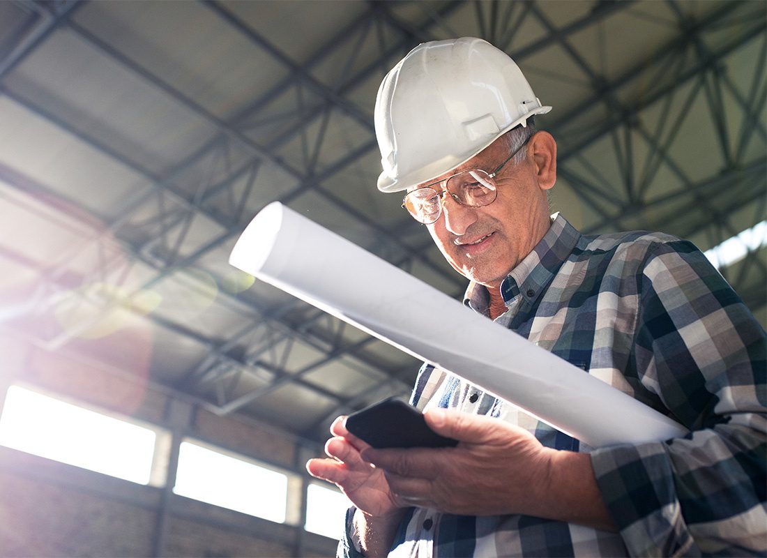 Read Our Reviews - Closeup Portrait of a Mature Senior Contractor at a Construction Jobsite Holding Blueprints While Looking at a Phone in his Hands